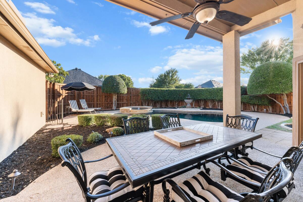 Covered patio with a ceiling fan and black-and-white cushioned dining set overlooking a landscaped backyard, swimming pool, stone fountain, trimmed hedges, and lounge chairs under a black umbrella on a sunny day.