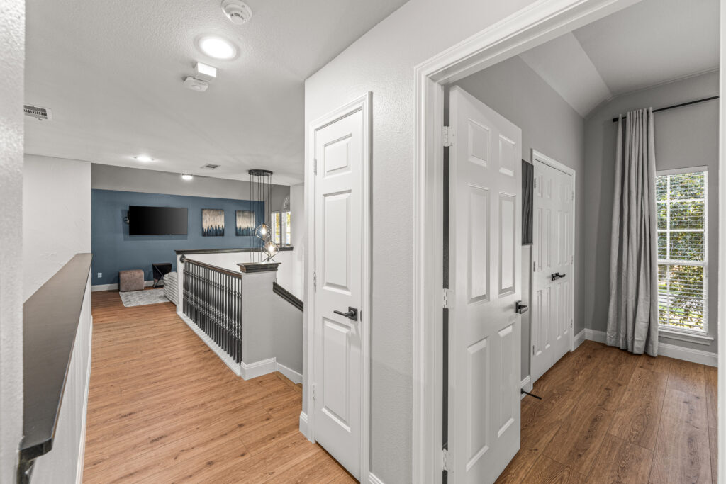 Upstairs hallway with wood flooring leading to a loft area that has a wall-mounted TV, artwork on a blue accent wall, and hanging light fixtures. A room with large windows and gray curtains is visible on the right.