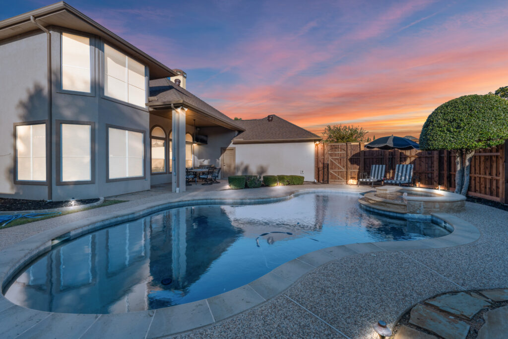 Backyard view at sunset featuring a swimming pool with curved edges, raised spa, and lounge chairs under a black umbrella. The two-story house has large windows, a covered patio with a dining set, and landscaped greenery along the wooden fence.